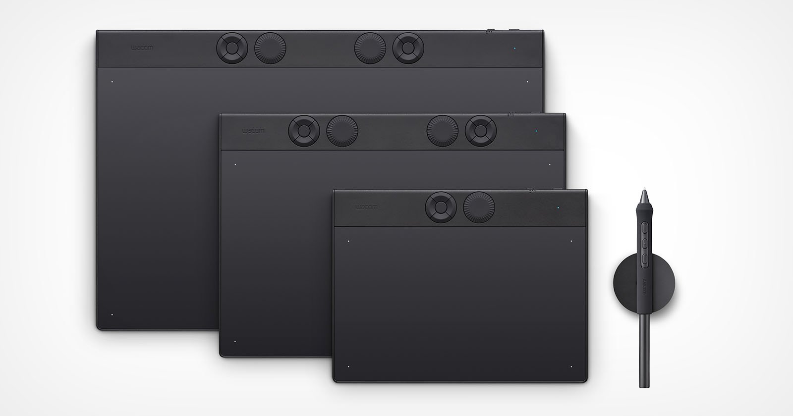 Three black drawing tablets of varying sizes are displayed vertically, each featuring buttons and dials. A stylus and a circular stylus holder are placed on the right. The background is plain white.