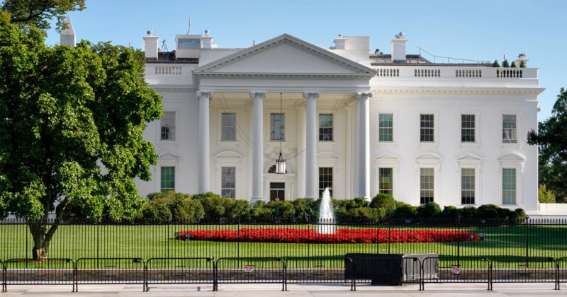 The image shows the front exterior of the White House, featuring a large white facade with columns and a triangular pediment. A manicured lawn with a circular flower bed and a small fountain is visible in the foreground, behind a black fence.