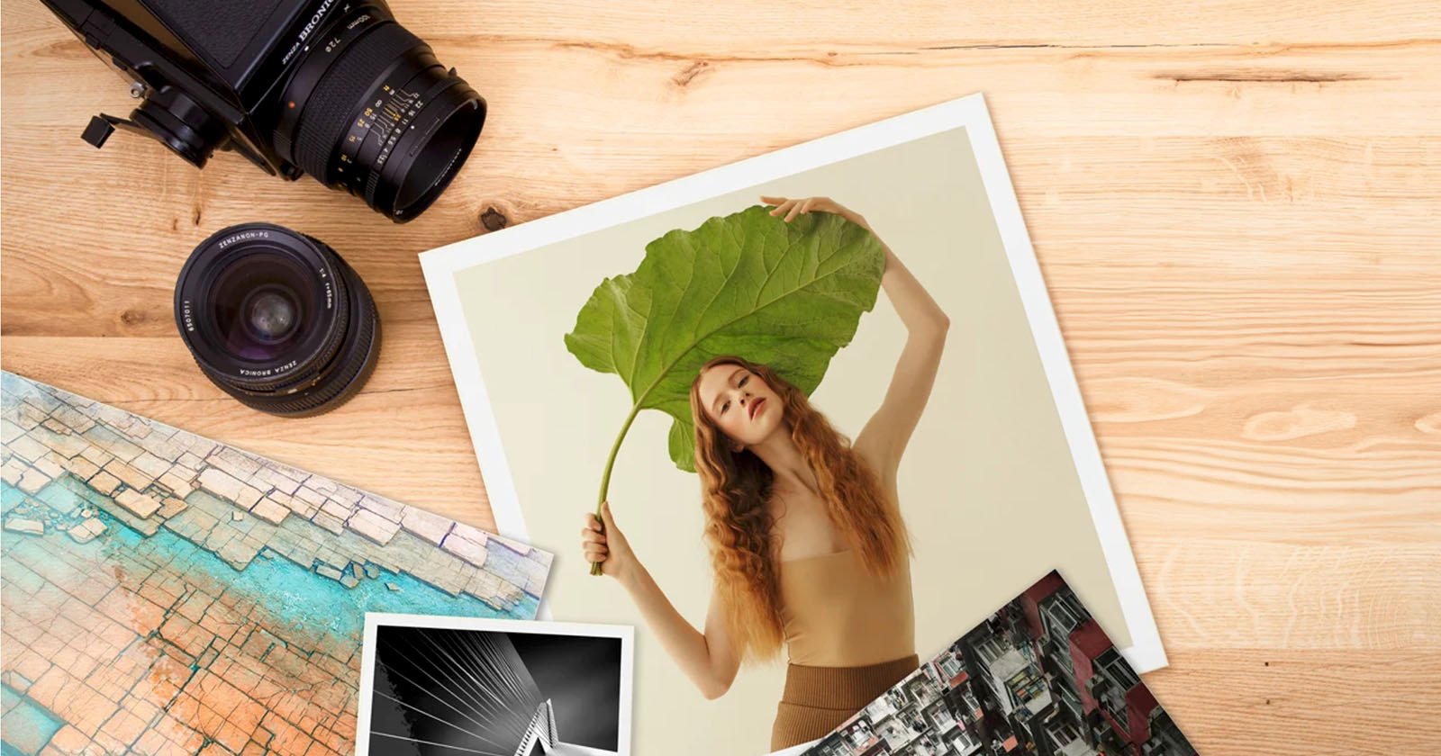 A wooden table with a camera, lens, map, and photographs. One photo shows a person holding a large leaf over their head. Another black-and-white photo depicts a suspension bridge, and the third photo shows a cityscape.