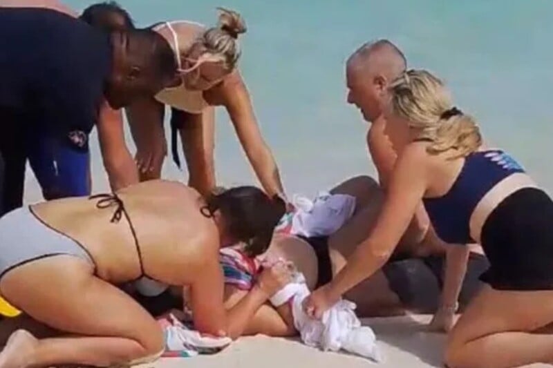 A group of people surround a person lying on a beach, assisting them. They appear to be offering help, with concern visible. The ocean is in the background, and the scene is set on sand under a clear sky.