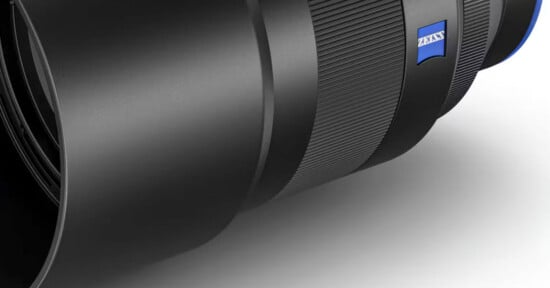 Close-up of a Zeiss camera lens showing the textured grip and blue Zeiss logo. The lens is positioned horizontally against a neutral background, highlighting its sleek black design.