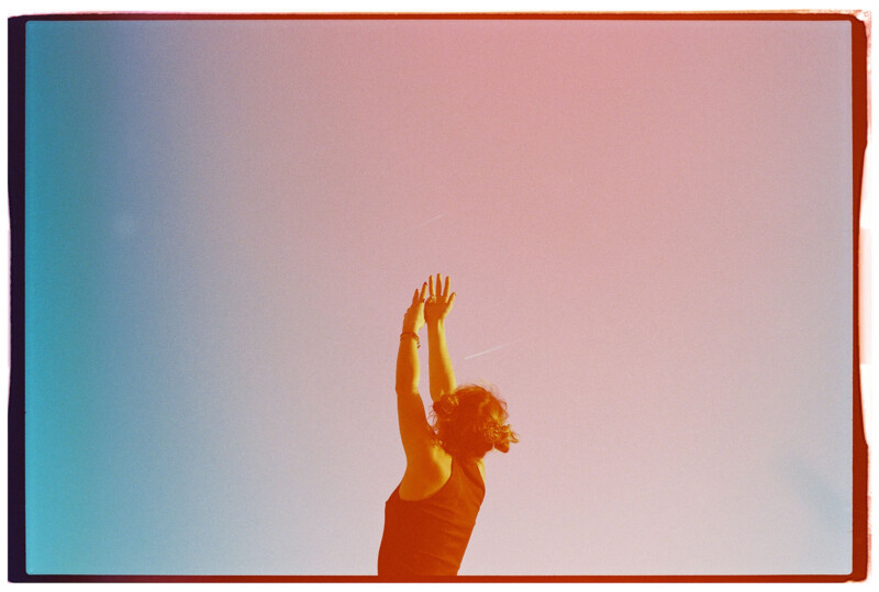 A person with short hair and a sleeveless top reaches upward with both hands against a sky backdrop. The image has a gradient overlay, transitioning from blue to red.