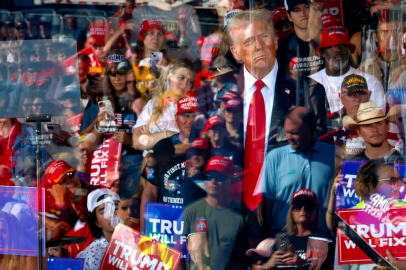 A group of people wearing red "Make America Great Again" hats, holding signs with slogans, and taking photos at a rally. The crowd's reflection overlays an image of a man in a suit, appearing as though he is behind glass.