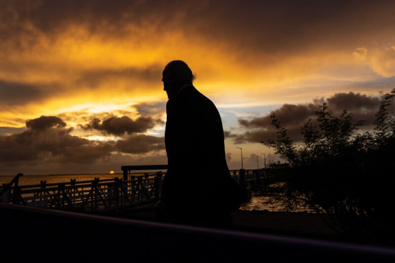 Silhouette of a person walking along a waterfront at sunset, with dramatic orange and yellow clouds in the sky. A pier is visible in the background, and there are bushes to the right.