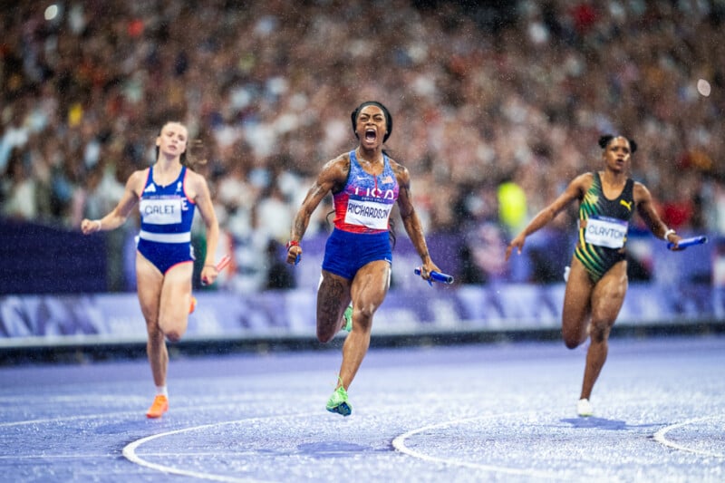 Three sprinters run on a wet track during a relay race. The central athlete, wearing a USA uniform, is shouting with excitement as they approach the finish line. A cheering crowd and blurred competitors are in the background.