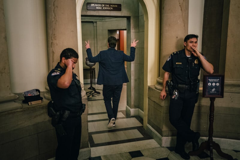 A person in a suit walks toward an office with a sign reading "Speaker of the House Mike Johnson," making a peace sign with both hands. Two uniformed officers stand nearby, one with a hand on their head and the other with a hand near their mouth.