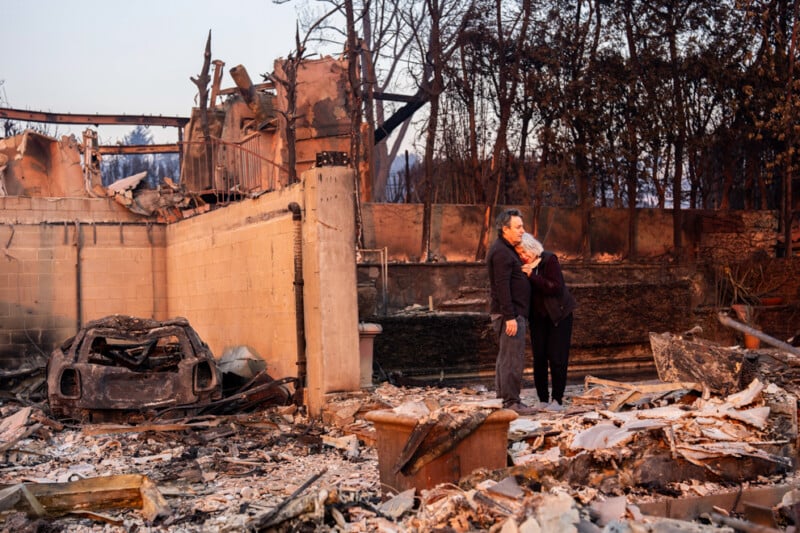 A man and woman stand amidst the charred remains of a house, with debris and a burnt car visible. The couple embraces, appearing emotional, surrounded by the ruins of what seems to be a severe fire. Burnt trees and skeletal remains of the structure are visible.