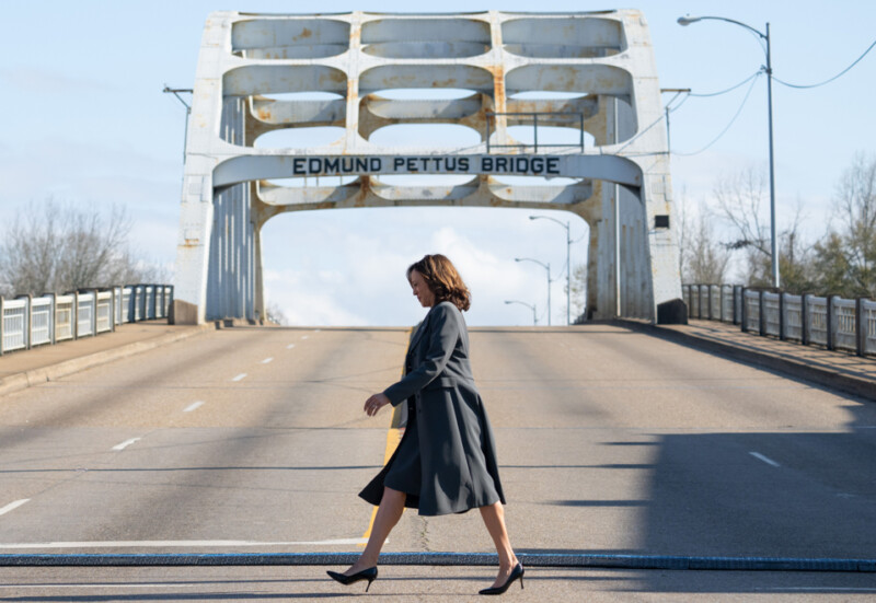 A person in a black coat and heels walks across the Edmund Pettus Bridge, with the sky visible in the background.