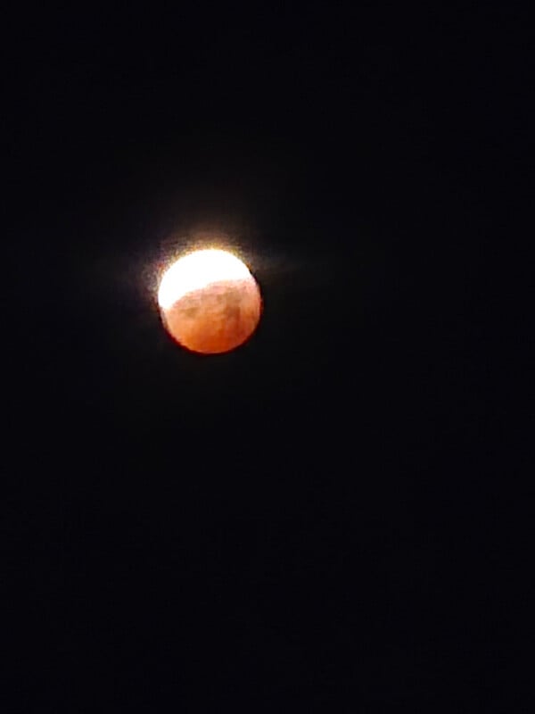 A partially eclipsed moon glows in the dark sky, with the Earth's shadow giving it a reddish tint on one side, and the other side shining brightly. The surrounding sky is black and featureless.