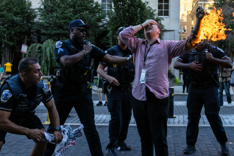 A man in a pink shirt with flames on his arm is surrounded by police officers on a city street. One officer reaches toward him with an outstretched arm. Other officers nearby are alert. Trees and a building are visible in the background.