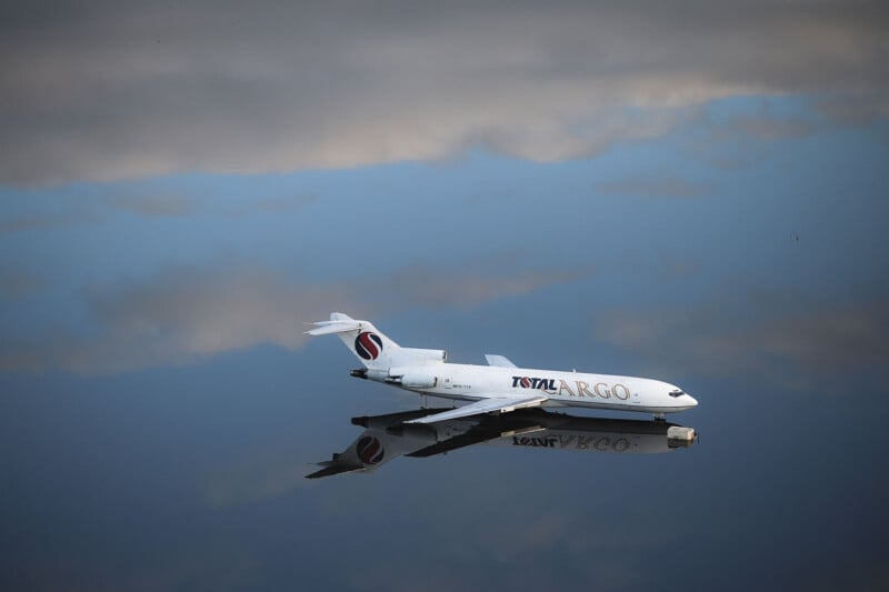 A white cargo plane lies partially submerged in a calm, reflective body of water. The reflection of the plane and scattered clouds is visible on the water's surface, creating a serene and surreal visual.