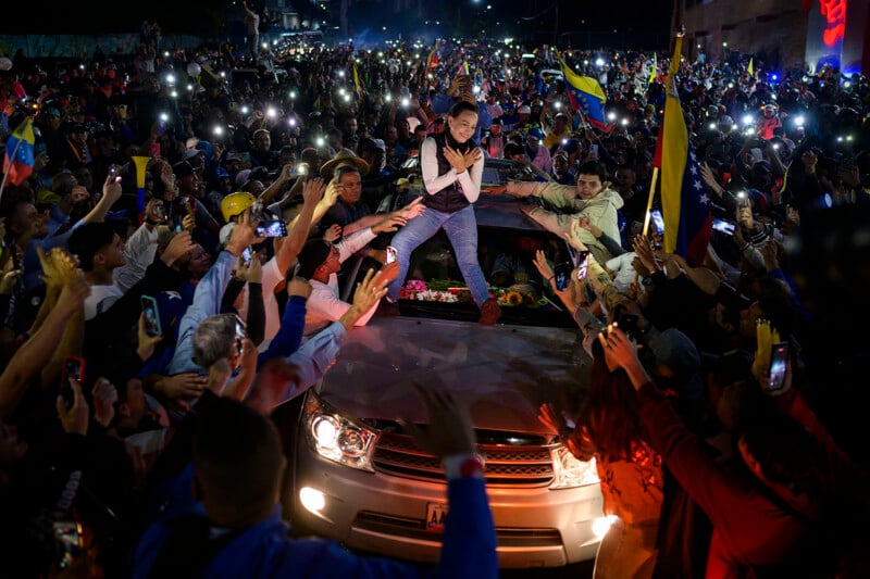 A woman stands on a car surrounded by a crowd at night. She has her hands crossed over her chest. People reach towards her while holding Venezuelan flags and smartphones, capturing the scene. The atmosphere is energetic and celebratory.