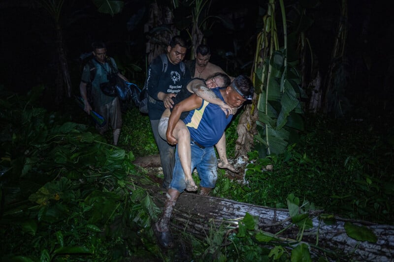 A group of men walk through a dense, dark jungle at night. One man carries another person on his back while others follow closely. The scene is dimly lit, highlighting the lush green foliage around them.
