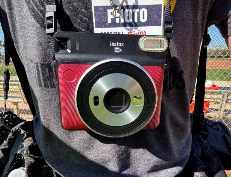 A person wearing a t-shirt has a large Instax SQ6 instant camera hanging around their neck. The camera is pink with a black and silver lens, and they also have a photo pass clipped to their shirt. A chain-link fence is visible in the background.