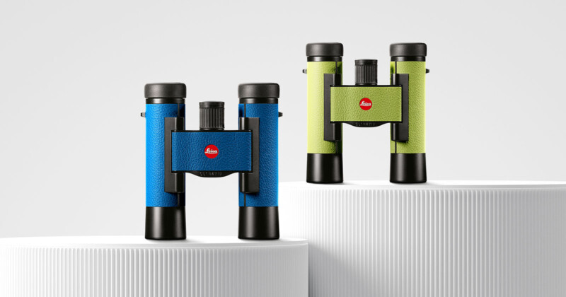 Two pairs of colorful binoculars with textured grips are displayed on white cylindrical pedestals. One pair is blue with black accents and the other is green with black accents, both featuring a red logo in the center.