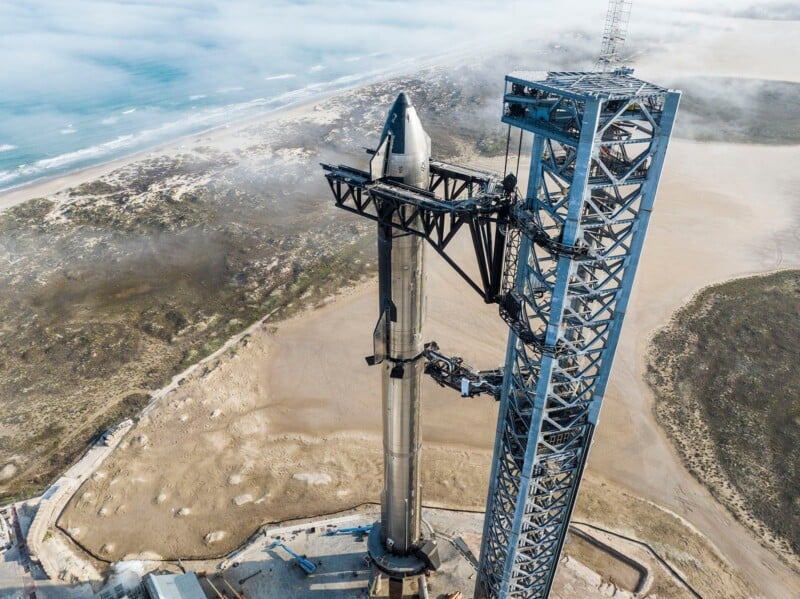 An aerial view of a rocket positioned vertically on a launch pad near the coastline. The ocean and sandy beach are visible in the background, with some clouds and mist surrounding the area.