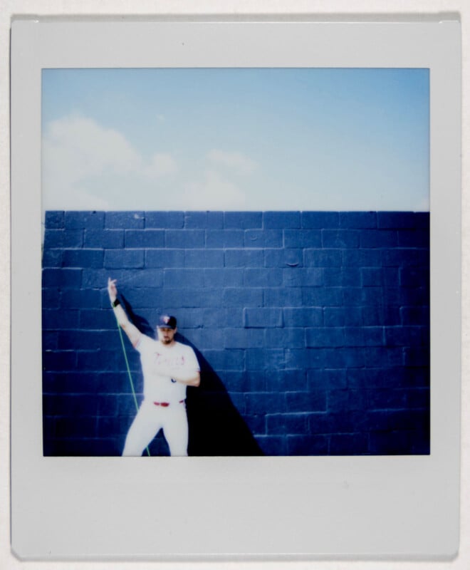 A person in a baseball uniform and cap is posing against a dark blue brick wall in a Polaroid photo. They are holding up a green exercise band with one arm. The sky is clear with a few clouds.