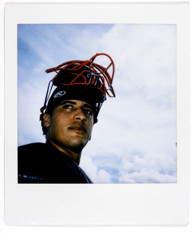 A baseball player wearing a black shirt and red catcher's helmet looks to the side. The background features a cloudy blue sky. The photo is in a Polaroid frame.