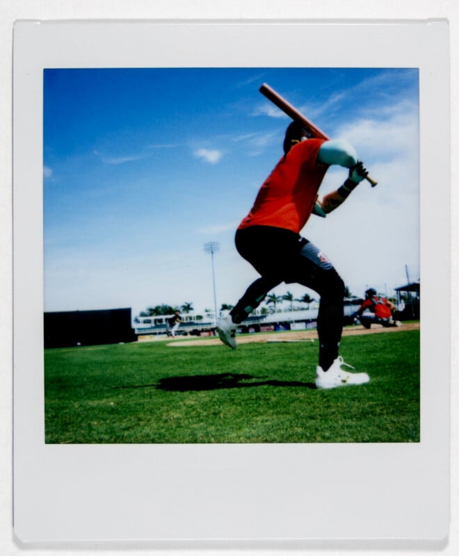 A baseball player in a red shirt and black pants prepares to swing a bat on a sunny day. The field is green with a clear blue sky and a few clouds. Another player and several spectators are visible in the distance.