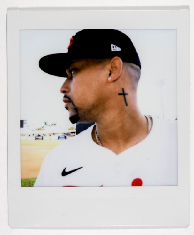 A man in a baseball uniform and cap looks to the side. He has a cross tattoo on his neck and a chain necklace. The background shows a baseball field under a clear sky.