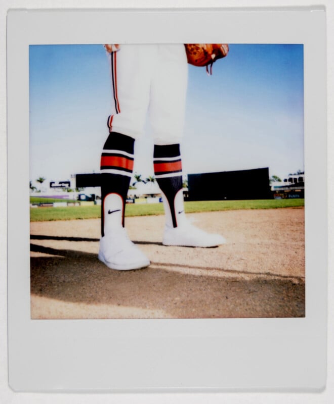 A Polaroid photo shows a baseball player from the thighs down standing on a field. The player wears a uniform with striped socks and white cleats. A baseball glove rests at their side. The sky is clear and the field is mostly empty in the background.