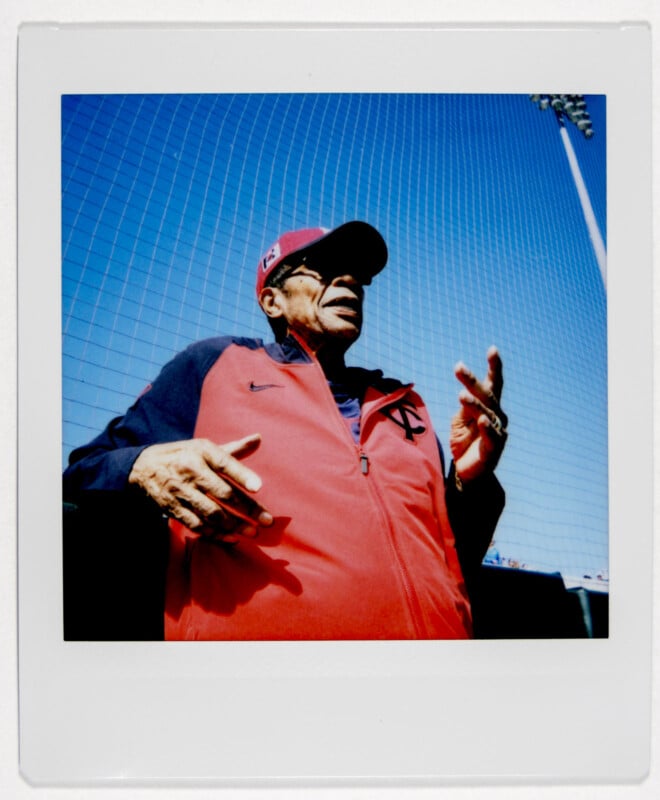 An individual wearing a red and blue baseball jacket and cap gestures with their hands. The sky is clear and a stadium light tower is visible in the background. The photo has a Polaroid style border.