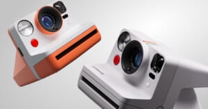 Two Polaroid instant cameras are shown against a light gray background. One camera is gray with an orange trim and the other is white with a rainbow decoration. Both have prominent lenses and viewfinders.