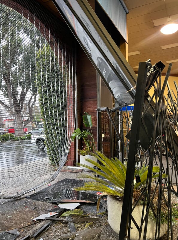 A damaged storefront with a partially collapsed metal security gate and framework. Broken glass and debris are scattered on the ground. Large potted plants and nearby trees are visible outside.