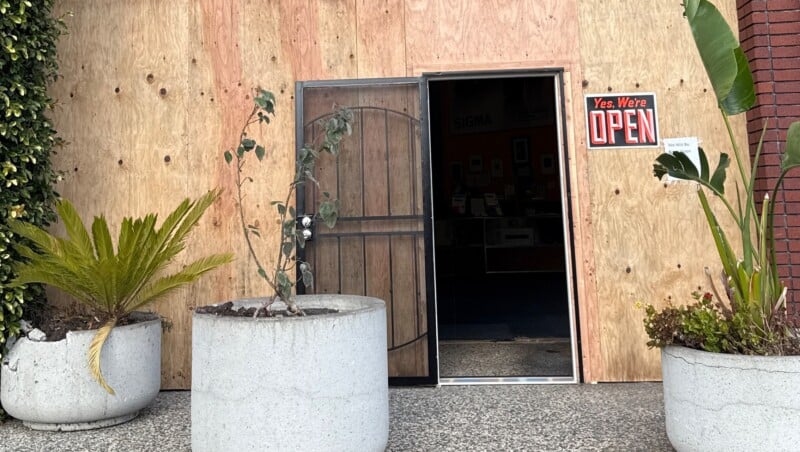 A storefront with a wooden facade and a partially open door displaying a bright red "Yes, We're Open" sign. Two large plants in gray pots are placed on either side of the entrance.