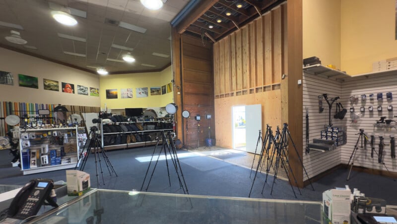 A photography equipment store interior with tripods displayed on the floor. Shelves on the right hold various camera accessories. Large photos and colorful backdrops line the left wall. A small open door lets in sunlight. A counter is visible in the foreground.