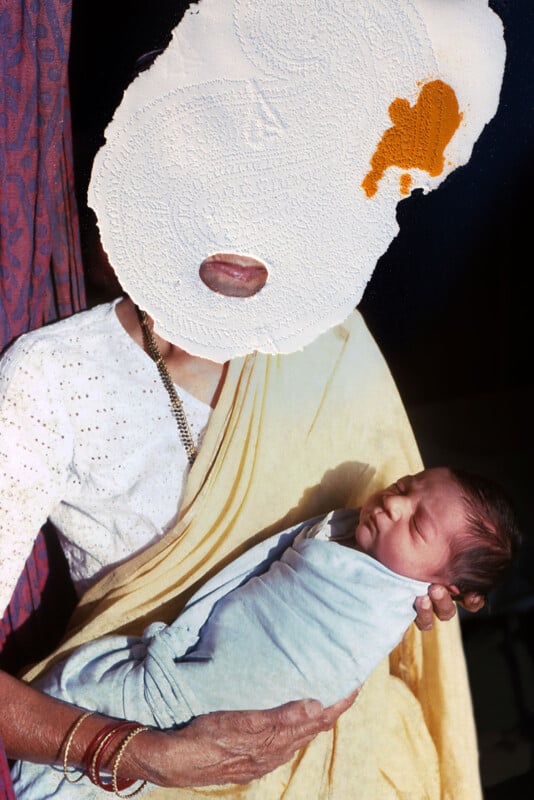 A person with a paper mask covering their face is holding a sleeping baby wrapped in a light fabric. The person is wearing a light yellow saree and gold bangles. The background is partially visible with a curtain.