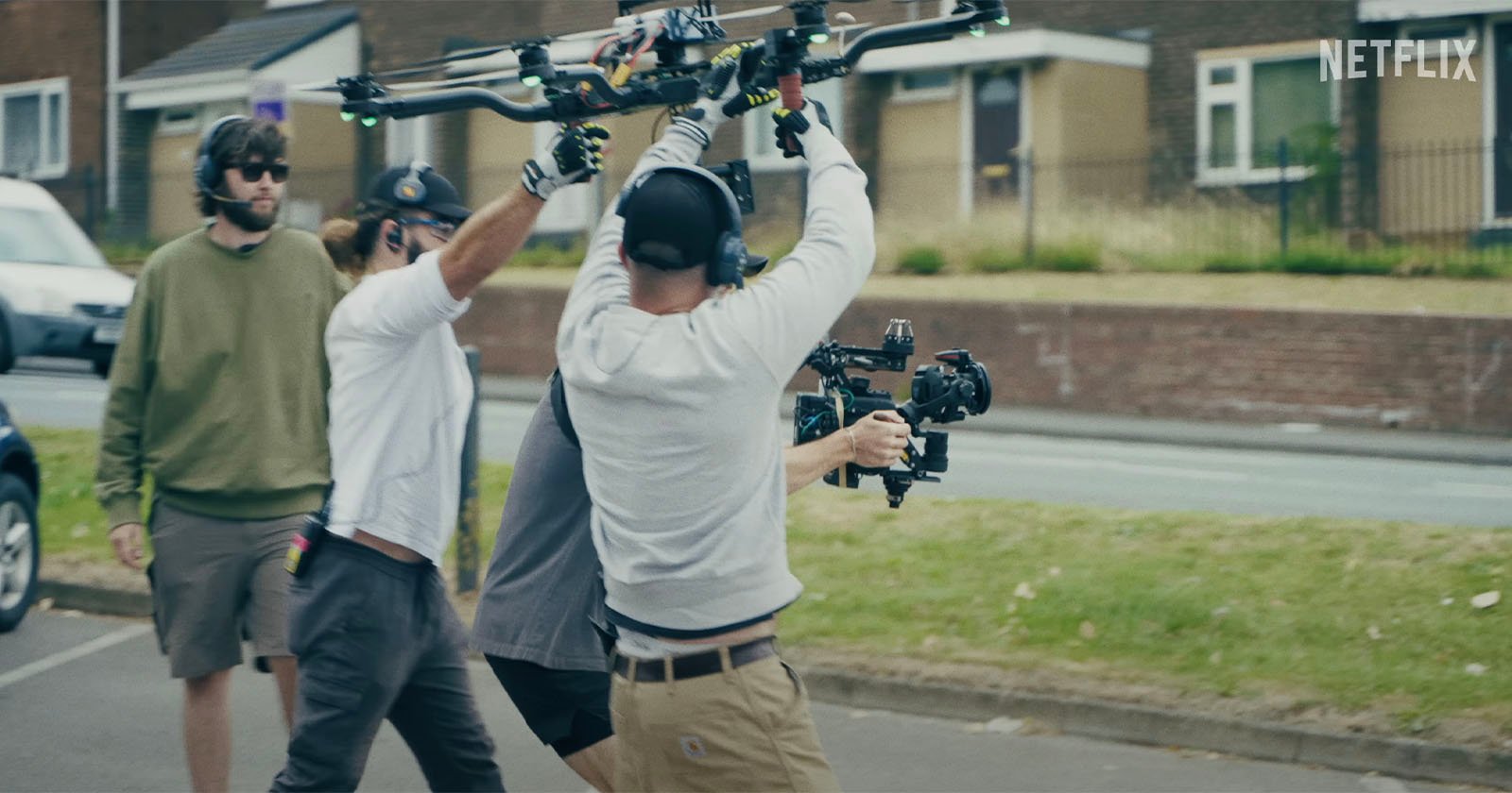 Crew members on a street work with a large drone, holding it overhead. They're setting up for filming. The drone is equipped with cameras. The setting includes grass and a building in the background.