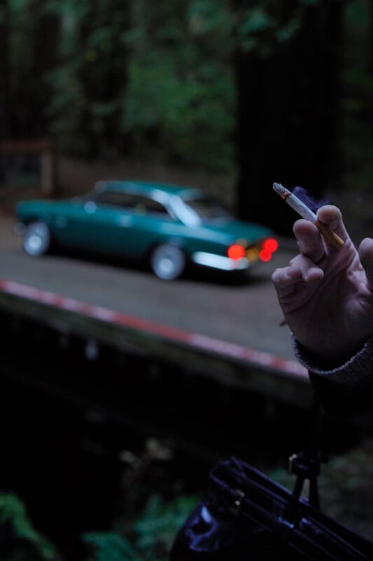 A hand holding a lit cigarette, partially in focus, appears in the foreground. In the background, there's a blurred view of a green vintage car parked by the roadside amidst lush greenery.