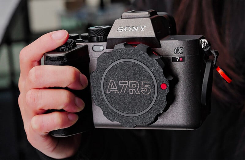 Close-up of a person holding a Sony A7R5 camera with a lens cap displaying "A7R5." The camera is black with various buttons and dials visible on the top.