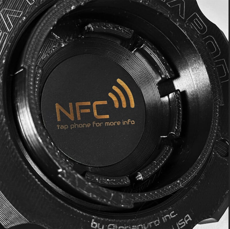 Close-up of a black cylindrical metal device with engravings, featuring a central circle displaying the NFC logo and the text "tap phone for more info" in yellow.