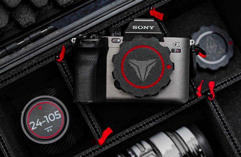 A Sony Alpha a7R III camera with a custom red and black lens cap is placed in a padded case. Next to it, there are camera lenses with red accents, including one labeled "24-105." The interior of the case is lined with black foam.