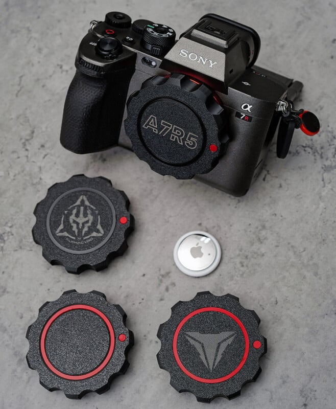 A Sony A7R5 camera is displayed on a textured gray surface. Below it are three custom lens caps with red and black designs, featuring different emblems. An Apple AirTag is also shown next to the caps.