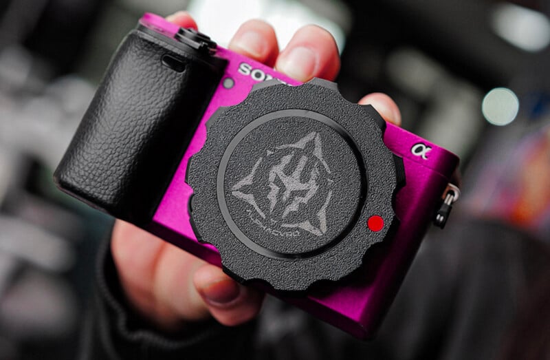 Close-up of a hand holding a purple Sony camera with a black grip and an artistic design on the lens cap. The camera appears compact, with a focus on its vibrant color and stylish details.