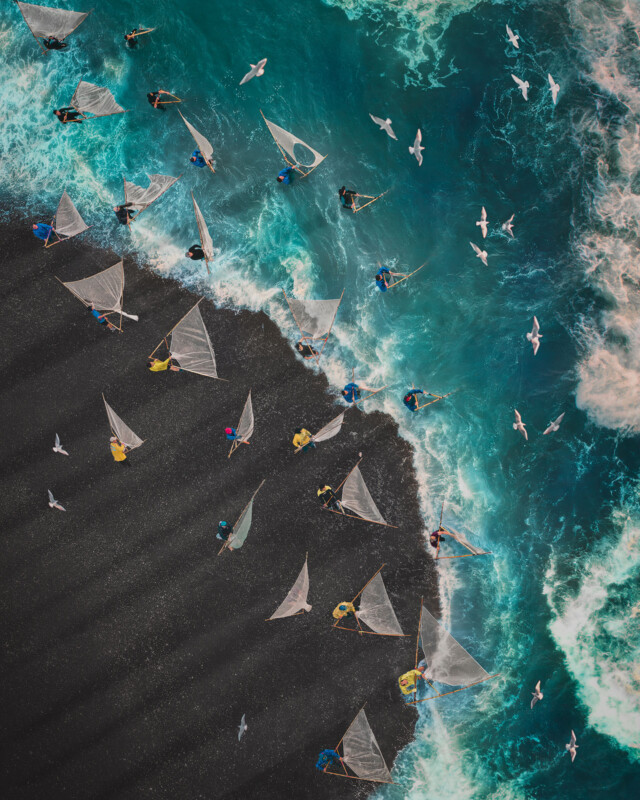 Aerial view of people with fishing nets standing on a shoreline where dark sand meets turquoise water. White waves crash nearby and several seagulls fly overhead. The scene is vibrant and dynamic.