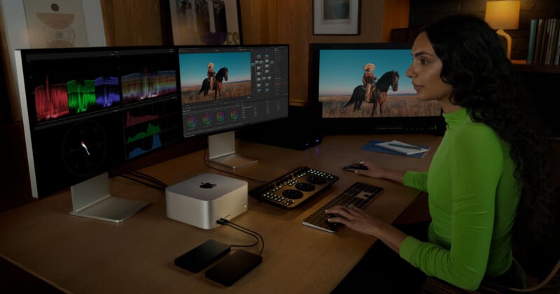 A person in a bright green shirt works at a desk with two large monitors displaying video editing software. A Mac mini and multiple peripherals are on the desk, and the room has wooden accents and low lighting.