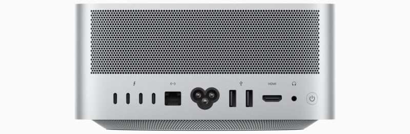 Rear view of a silver computer with perforated metal design. Visible ports include Thunderbolt, USB-C, USB-A, HDMI, Ethernet, and a power button on the right side. A power connector is centrally located.