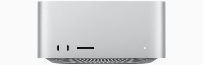 Front view of a compact, silver computer unit. It features a minimalist design with a smooth surface. The lower section is perforated for ventilation, while the front has two narrow vertical slots and a circular power indicator.