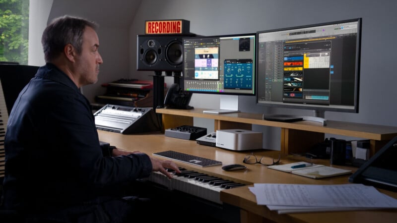 A person sits at a keyboard in a music studio, facing two computer monitors displaying music production software. A "RECORDING" sign and speakers are on the desk. Papers and electronic devices are on the wooden desk.