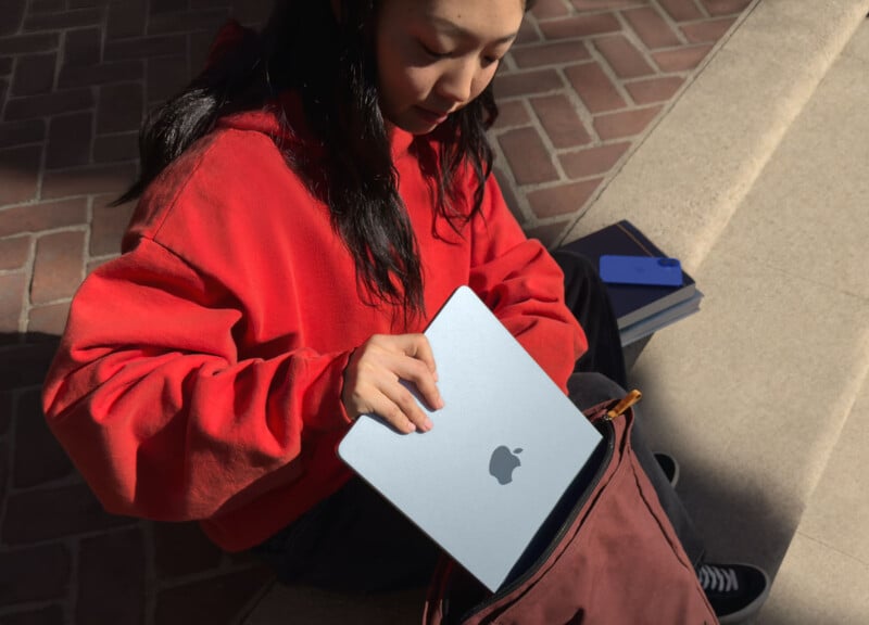 A person in a red sweatshirt sits on steps and takes a silver laptop with an apple logo out of a red bag. A couple of notebooks and a blue phone are on the step beside them.