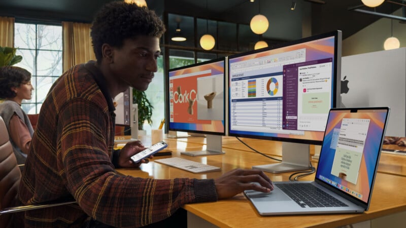 A person is working at a desk with multiple screens. They have one hand on a laptop and are holding a smartphone in the other. The monitors display various applications. Another person is in the background in an office setting.