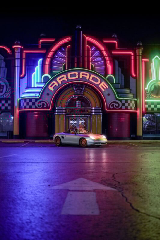 A silver sports car is parked in front of a brightly lit arcade. The building features colorful neon lights in pink, blue, and green, with large letters spelling "ARCADE" above the entrance. The ground reflects the vibrant lights.