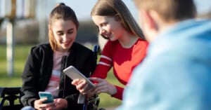 Two young women sitting outdoors, smiling and looking at their smartphones. One is in a wheelchair, and they appear to be sharing something on their screens. A person in a blue hoodie is blurred in the foreground.