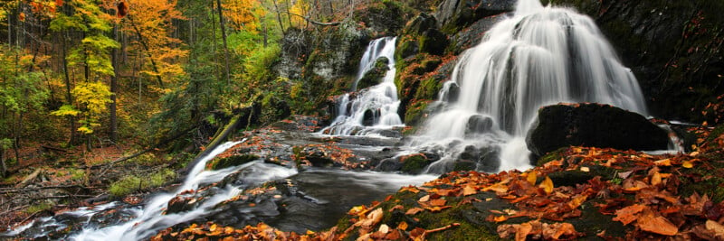 A picturesque waterfall cascades over rocks in a forest during autumn. The scene is filled with vibrant fall foliage in shades of orange, yellow, and green, blanketing the forest floor and surrounding the flowing water.