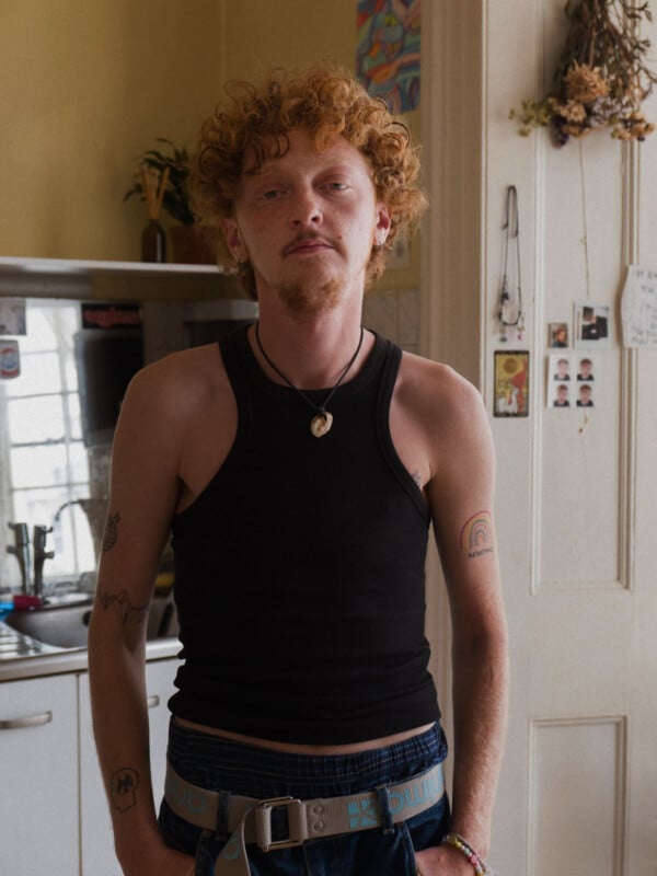 A person with curly red hair wearing a black tank top stands in a kitchen. They have several tattoos on their arms and a necklace. The kitchen has a sink, plants, and items hanging on the wall. The mood appears casual and relaxed.