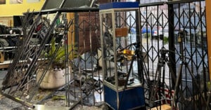 A store interior with metal security grilles bent and glass cases damaged, displaying signs of a break-in. Shattered glass covers the floor alongside fallen plants and camera equipment.